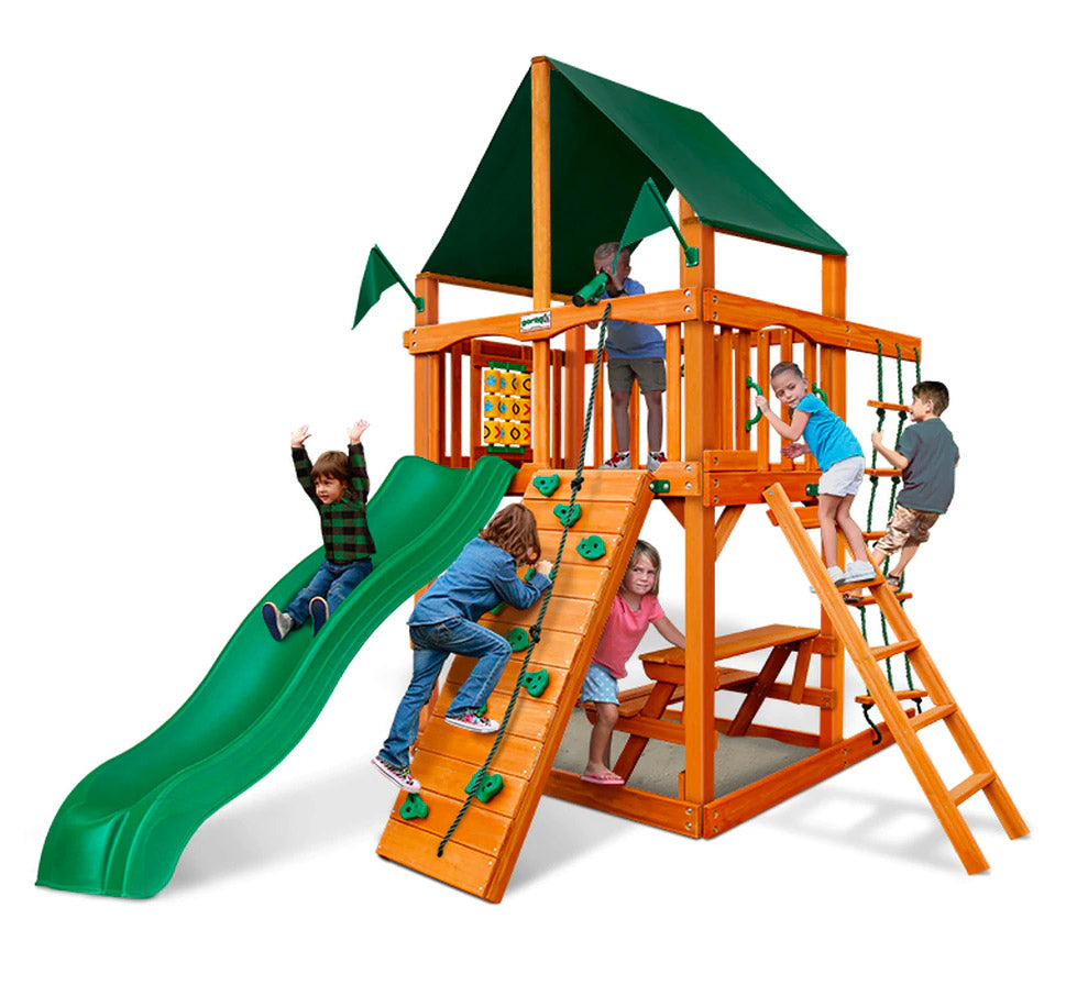Wooden playset with green Sunbrella canopy, rock climbing wall, slide, swings, picnic table, and tic-tac-toe panel with children playing