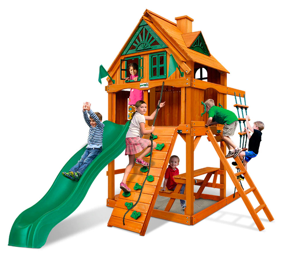 Wooden playset with green slide, rock climbing wall, rope ladder, picnic table, and children playing outdoors