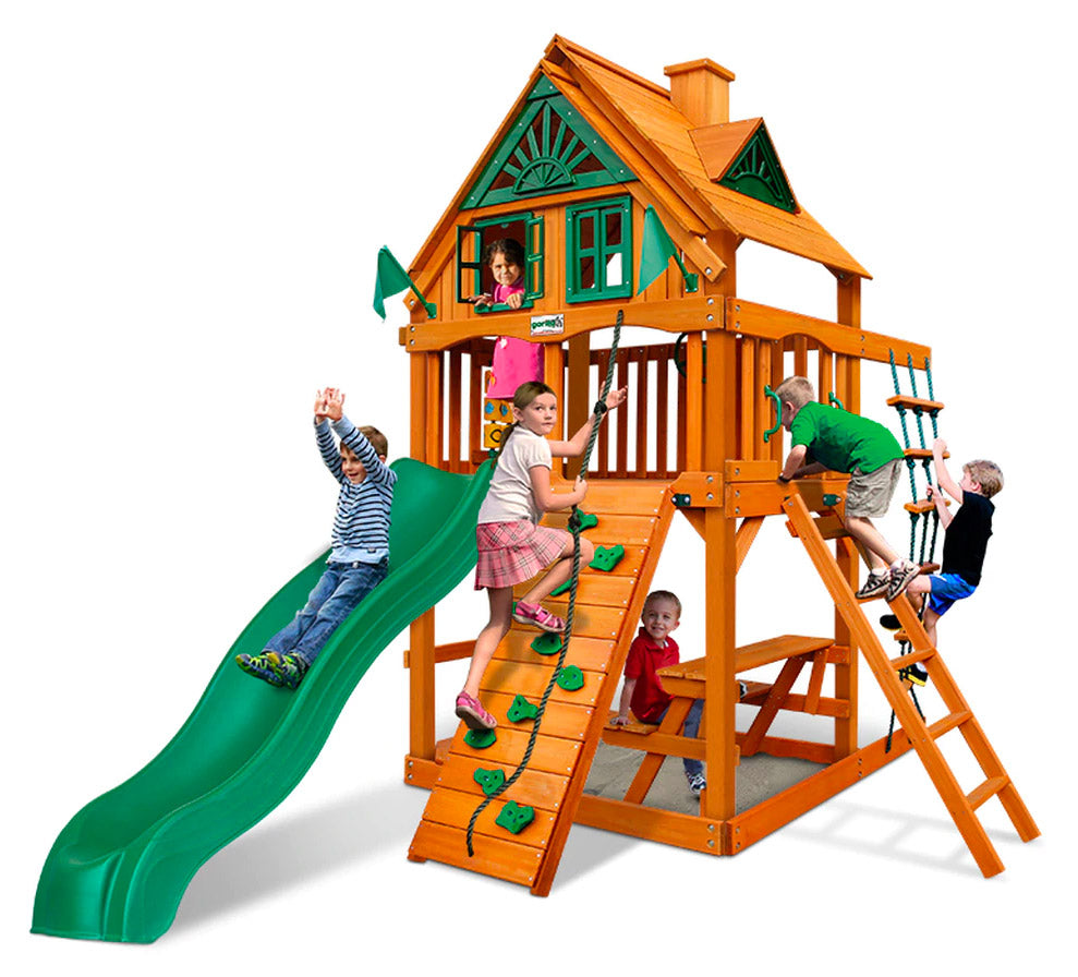 Wooden playset with green slide, climbing wall, rope ladder, picnic table, and upper-level playhouse with shutters and children playing