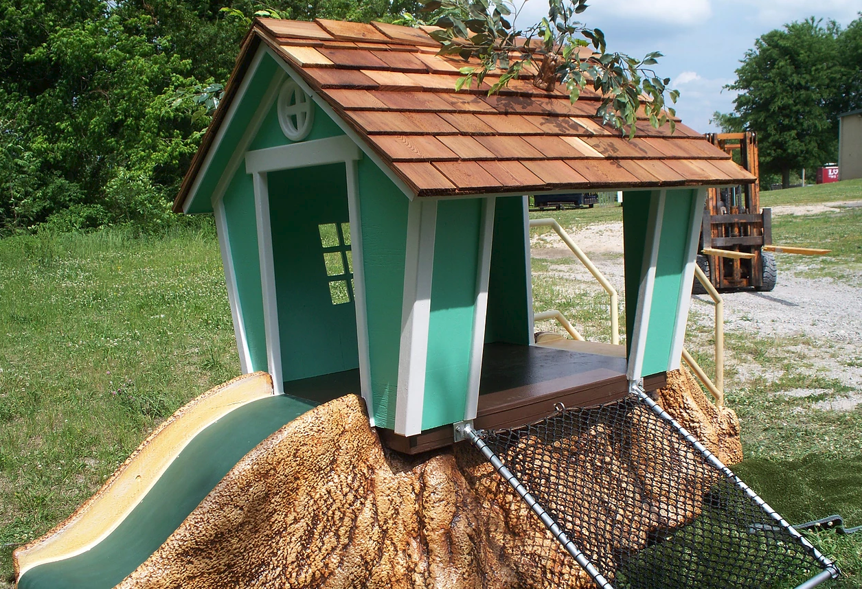 Custom-themed water slide with green and white playhouse, brown shingled roof, textured rock base, and climbing net.