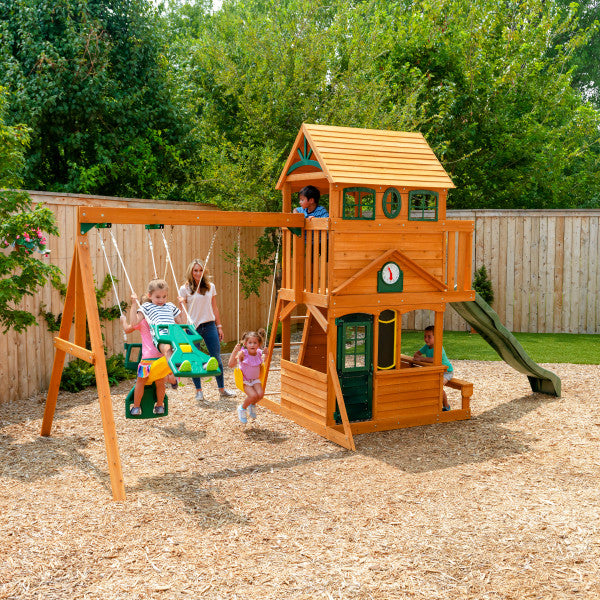 Two-story wooden playset with swings, rock wall, slide, clubhouse, play clock, and children playing outdoors