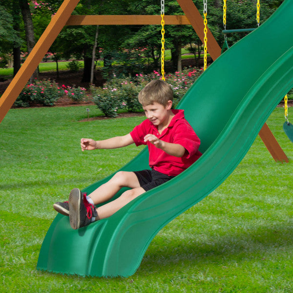 Child sliding down a green alpine wave slide attached to a cedar wooden swing set with yellow chains on grass