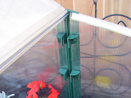 Green double cold frame with clear polycarbonate panels and red flowers inside, featuring adjustable roof windows and rounded edges