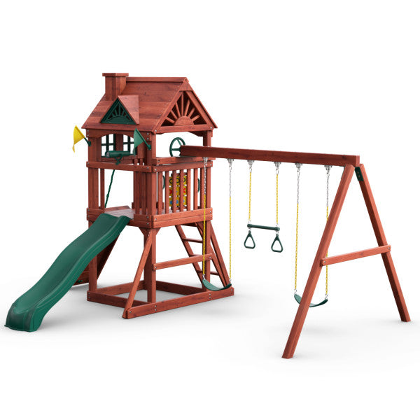 Redwood-finished wooden playset with green wave slide, two belt swings, trapeze bar, climbing ladder, and upper clubhouse with chimney and shutters