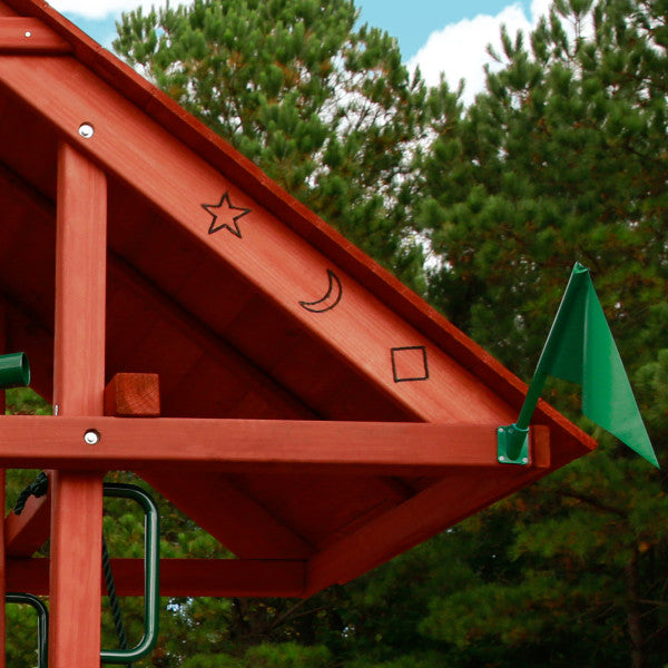 Redwood-stained wooden playset roof with sunburst cutouts, green flag, and metal hardware among pine trees