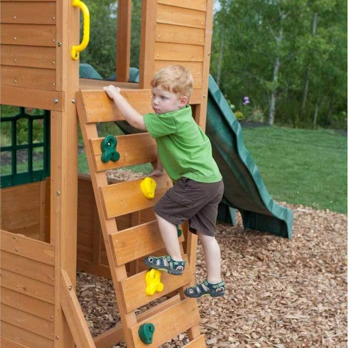 Child climbing colorful wooden rock wall on cedar playset with green slide and wood chip ground cover