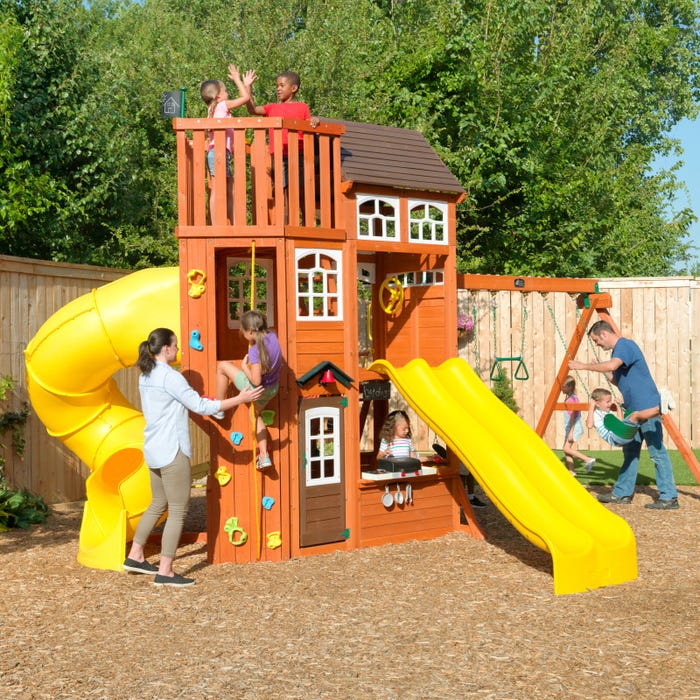 Wooden multi-level playset with yellow double slide, twist tube slide, climbing wall, swings, and children playing outdoors