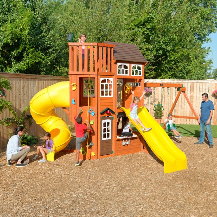 Wooden playset with two yellow slides, climbing wall, rope ladder, upper deck with windows, and children playing outdoors