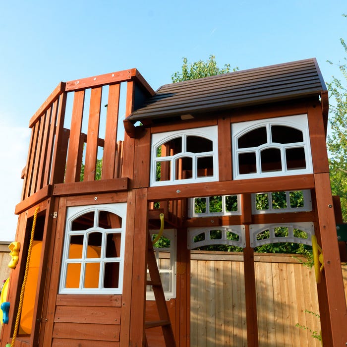 Wooden two-story playset with white-framed windows, shiplap roof, climbing ladder, and backyard setting