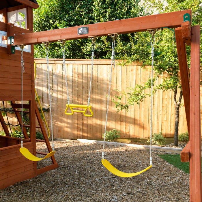 Wooden multi-level play structure with two yellow belt swings and a yellow trapeze bar in a backyard setting