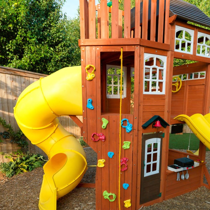 Wooden playset with colorful climbing holds, yellow twist tube slide, windowed clubhouse, and play kitchen with utensils