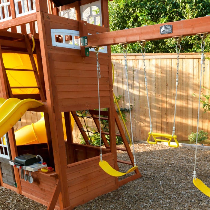 Wooden playset with yellow slides, swings, climbing ladder, and play BBQ area in a backyard setting
