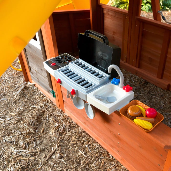 Wooden children's playset with attached toy BBQ grill, sink, plastic play food, and wood chip ground surface.