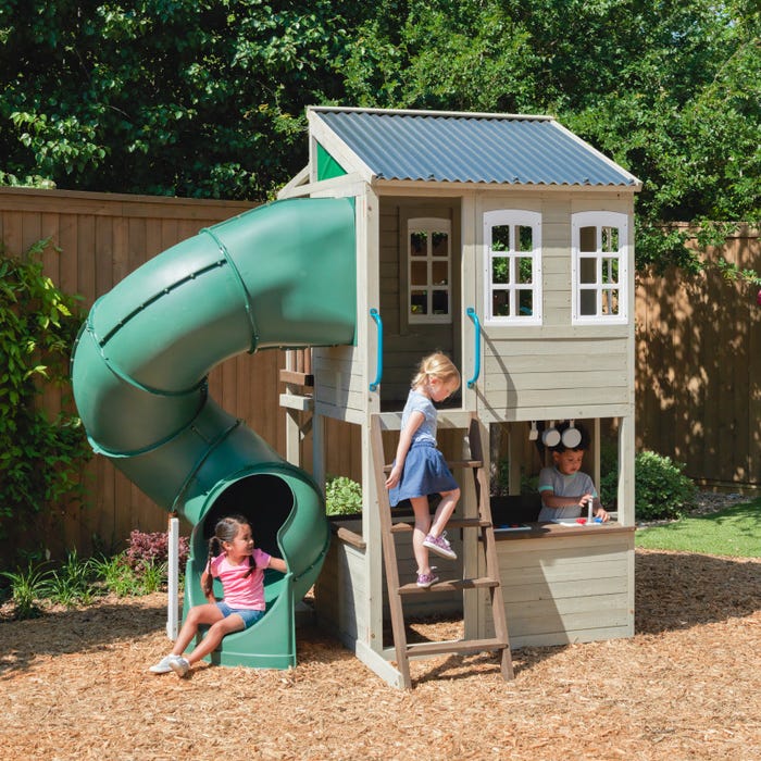 Two-story wooden playhouse with green twisty slide, ladder access, multiple windows, and children playing outdoors