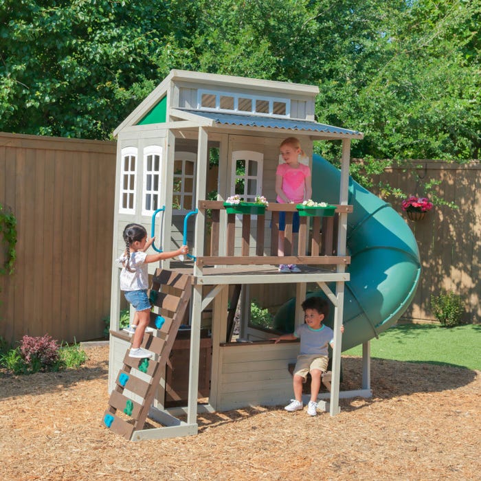 Two-story wooden playhouse with twisty slide, rock climbing wall, flower boxes, and children playing outdoors