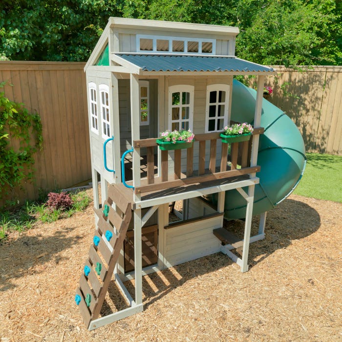 Two-story wooden playhouse with twisty tube slide, rock climbing wall, open deck with flower boxes, and ladder access in backyard setting
