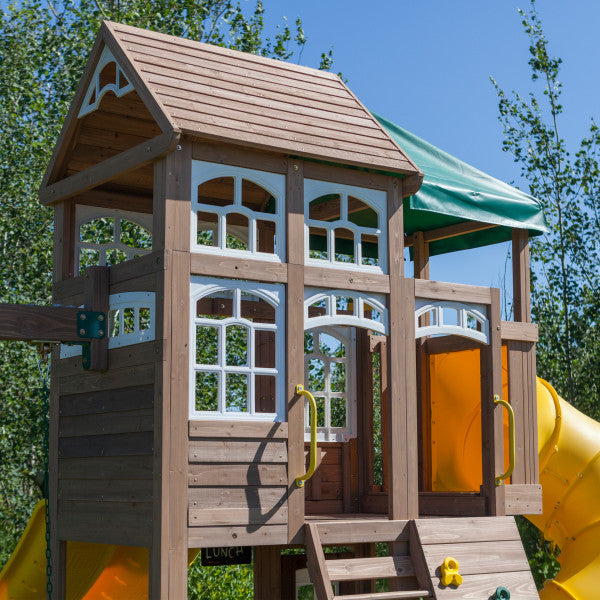 Two-level wooden playset with decorative windows, green canopy, yellow twist slide, climbing ladder, and swings.