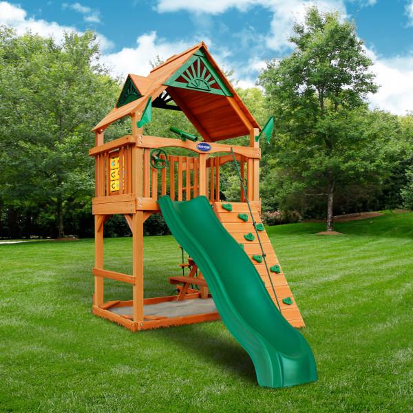 Cedar wooden playset with green slide, climbing wall, tic-tac-toe panel, and roofed upper deck in grassy backyard