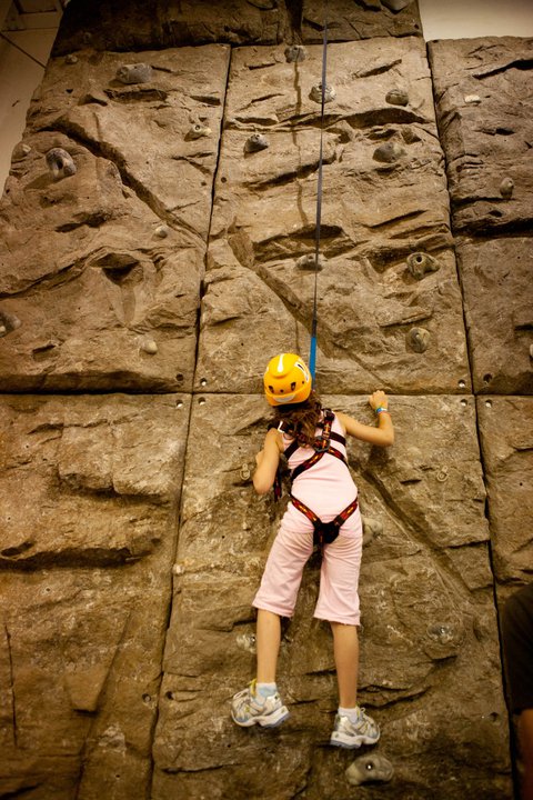Child wearing safety harness and helmet climbing a textured modular climbing panel with colorful handholds