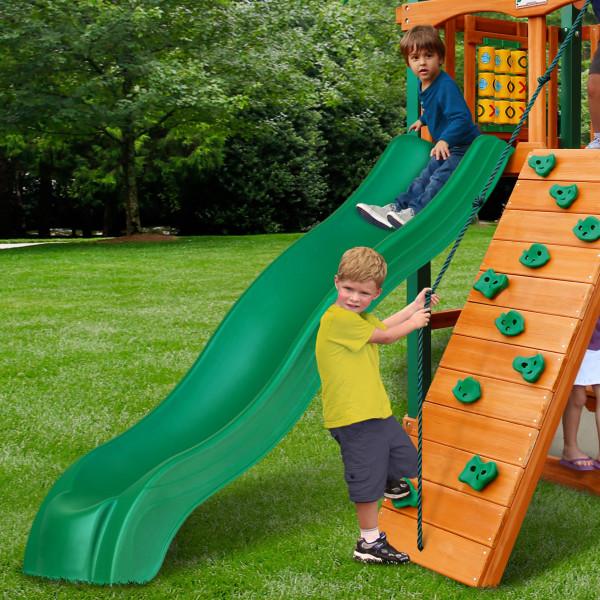 Wooden playset with green alpine wave slide, rock climbing wall with rope, tic-tac-toe panel, and children playing outdoors