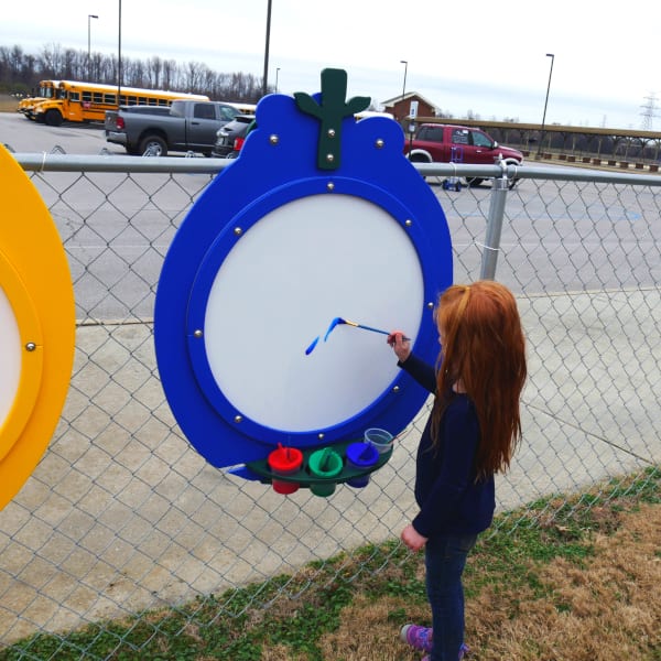 Child painting on blue fruit-shaped outdoor art panel with attached paint cups on chain-link fence