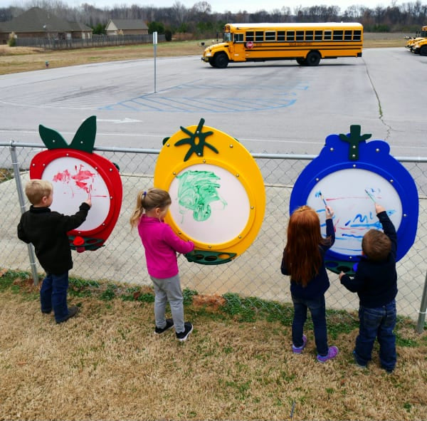 Children drawing on colorful fruit-shaped art panels attached to a fence in an outdoor play area near a school bus