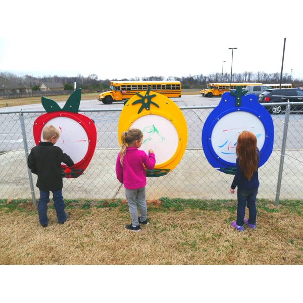 Three children drawing on colorful fruit-shaped outdoor art panels mounted on a playground fence, promoting creative play and collaboration.