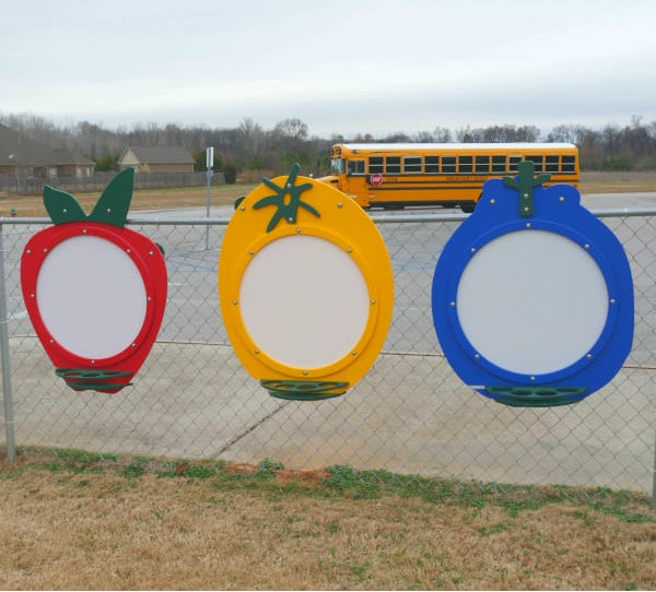 Colorful fruit-shaped art panels in red, yellow, and blue mounted on a fence for outdoor children's creative play.