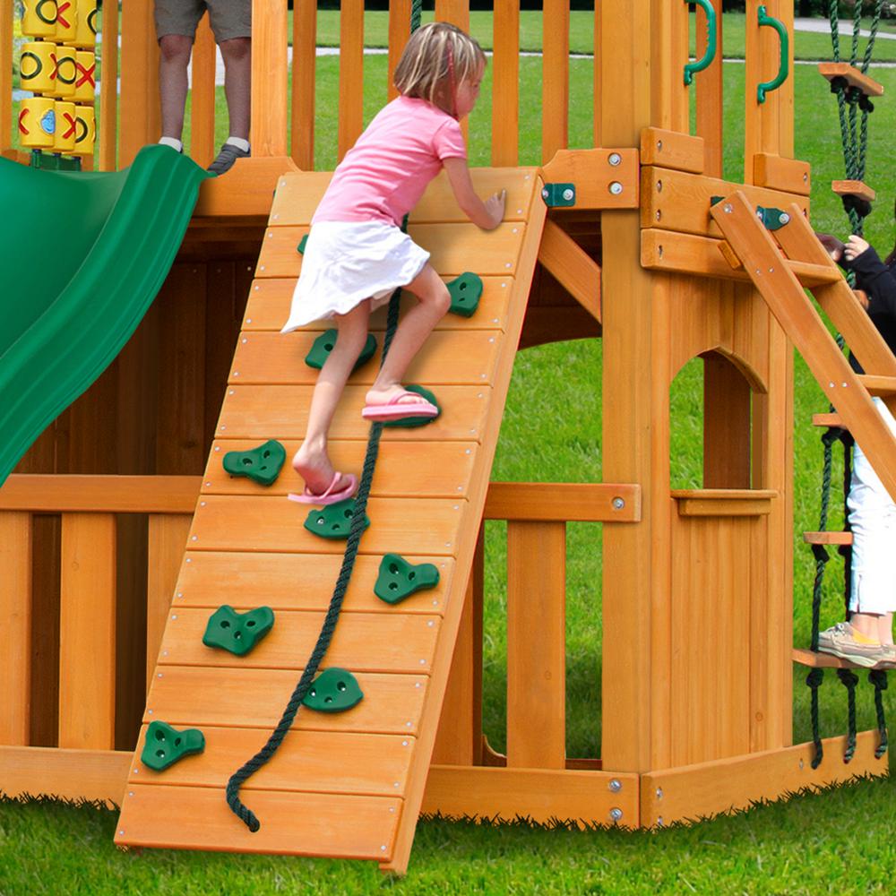 Child climbing amber-finished cedar wooden swing set rock wall with green handholds and climbing rope