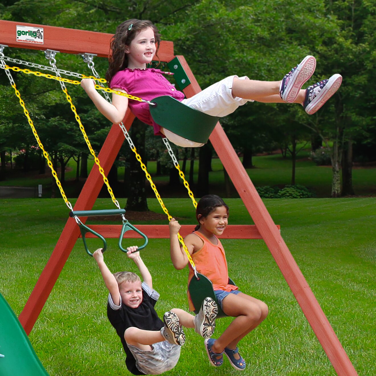 Three children playing on a wooden swing set with green belt swings and yellow chains in a grassy backyard