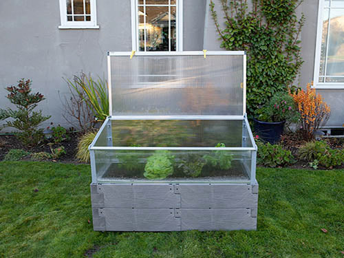 Gray poly timber textured raised garden bed with clear polycarbonate cold frame cover open, containing green leafy plants.