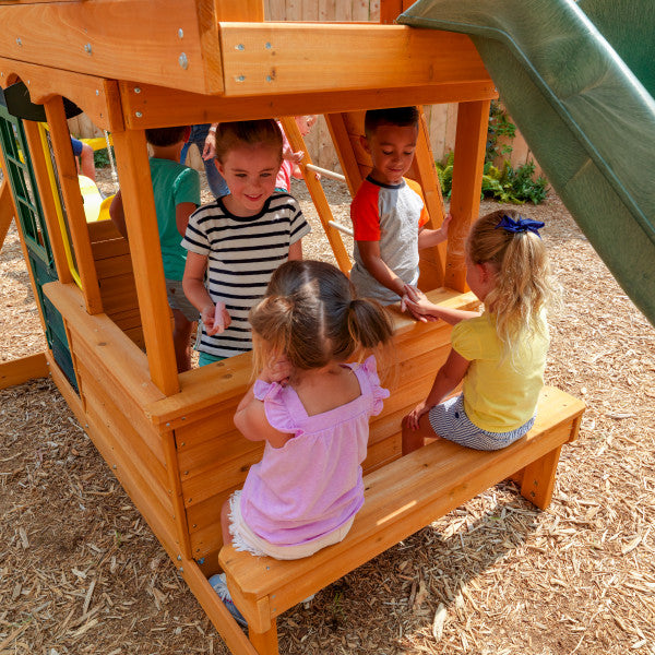 Children playing on a wooden playhouse with bench seating, green high rail wave slide, and wood chip ground cover