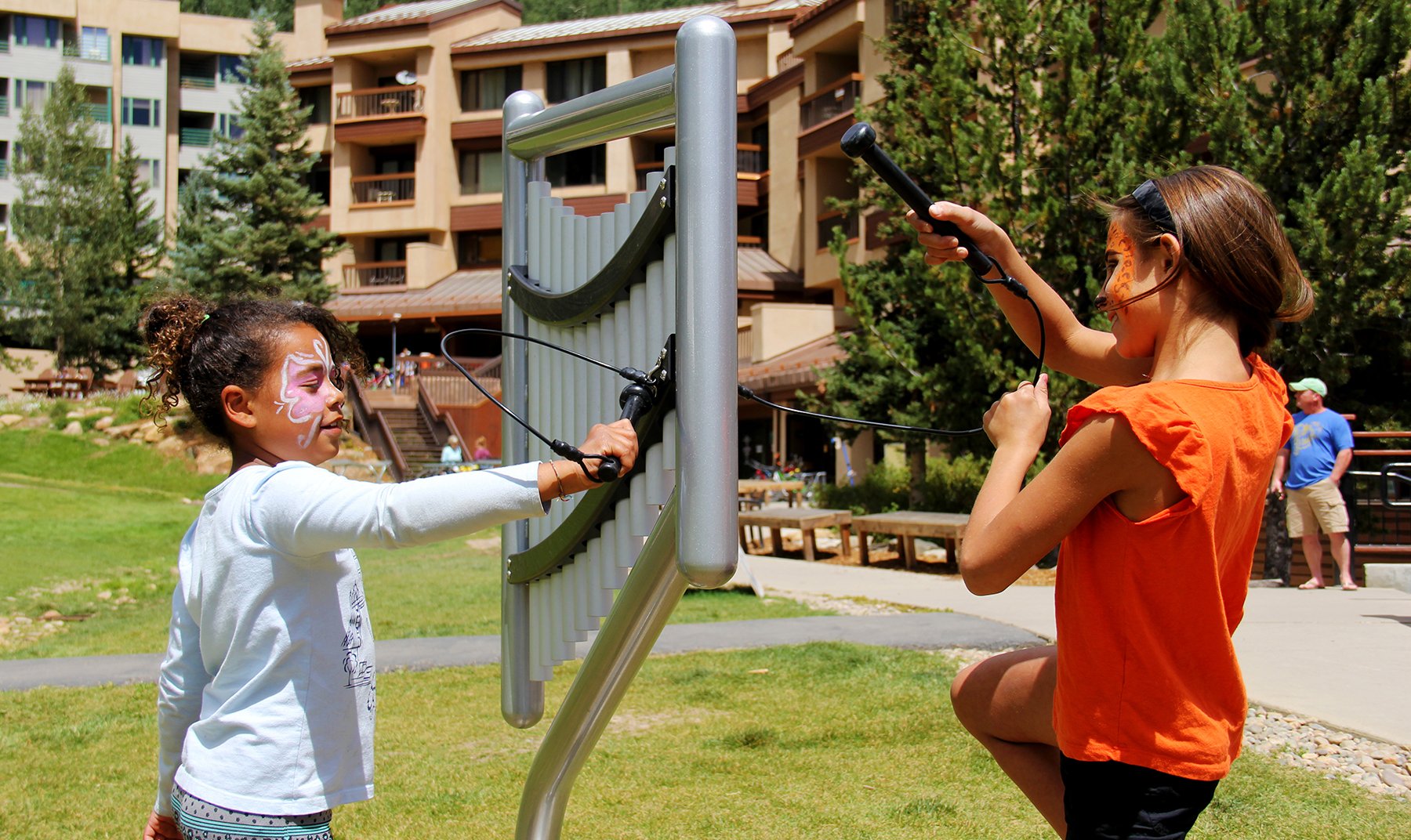 Two children with face paint playing a silver outdoor harp-style musical chime instrument in a park setting