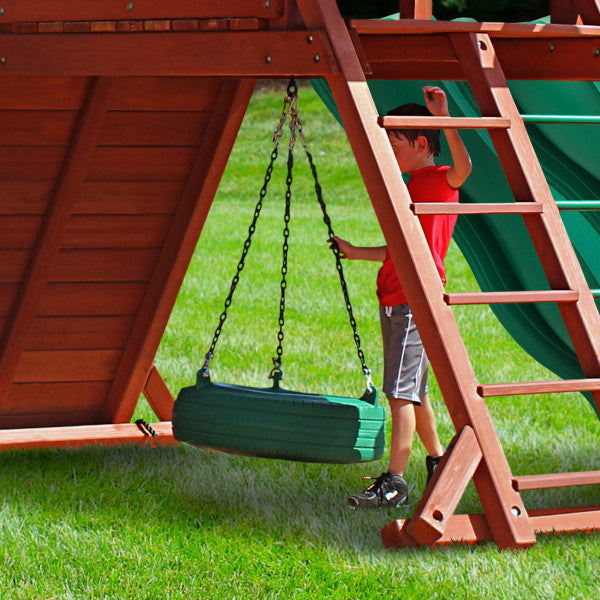 Cedar wood swing set with green tire swing, ladder, and green slides on grass with child in red shirt