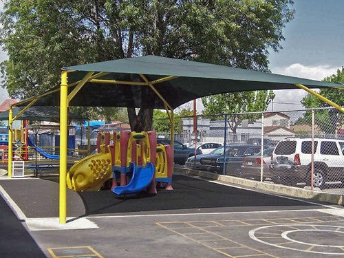 Rectangular hip roof shade structure with four yellow posts covering playground equipment and picnic area, UV-blocking fabric.