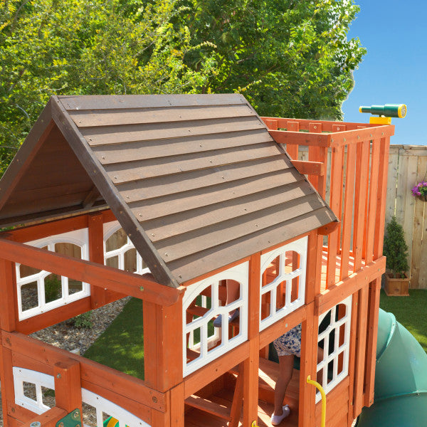 Multi-level wooden playset with shiplap roof, white-framed windows, green tube slide, yellow telescope, and double wave slide