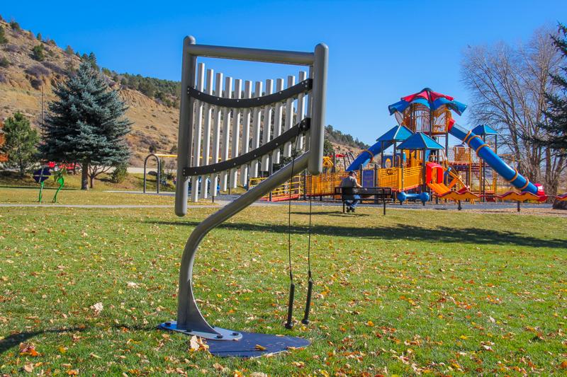 Outdoor harp-style musical instrument with 11 anodized aluminum chimes and mallets on grassy lawn near colorful playground equipment