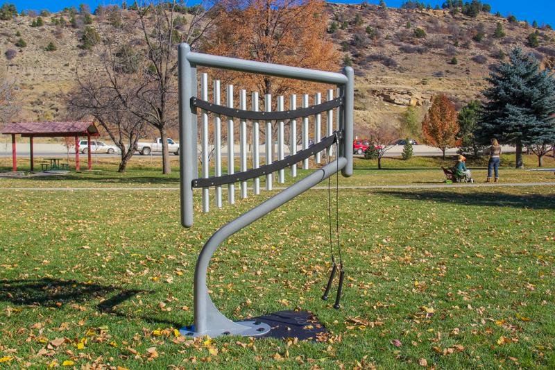 Outdoor harp musical instrument with 11 anodized aluminum chimes on a curved gray frame in a park setting