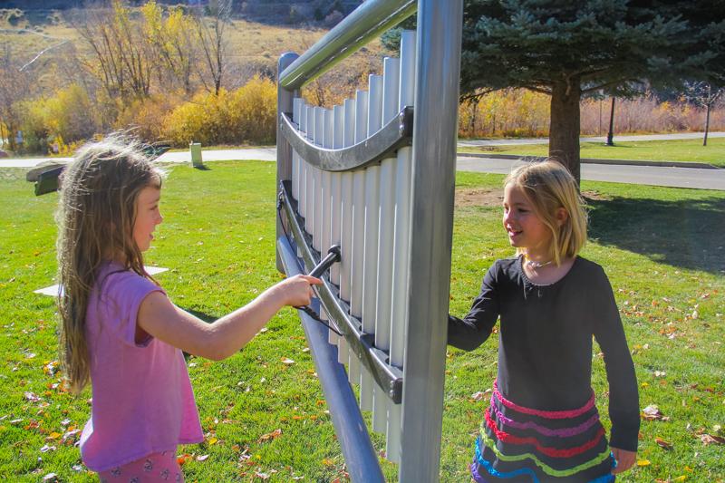 Two children playing a large outdoor harp-style musical instrument with anodized aluminum chimes in a sunny park setting