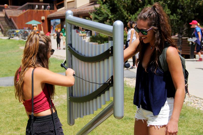 Two young women playing a silver outdoor musical chime instrument with anodized aluminum tubes in a sunny park setting