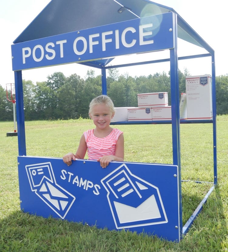 Child playing in a blue outdoor playhouse with mail-themed panels and postal package props on grass