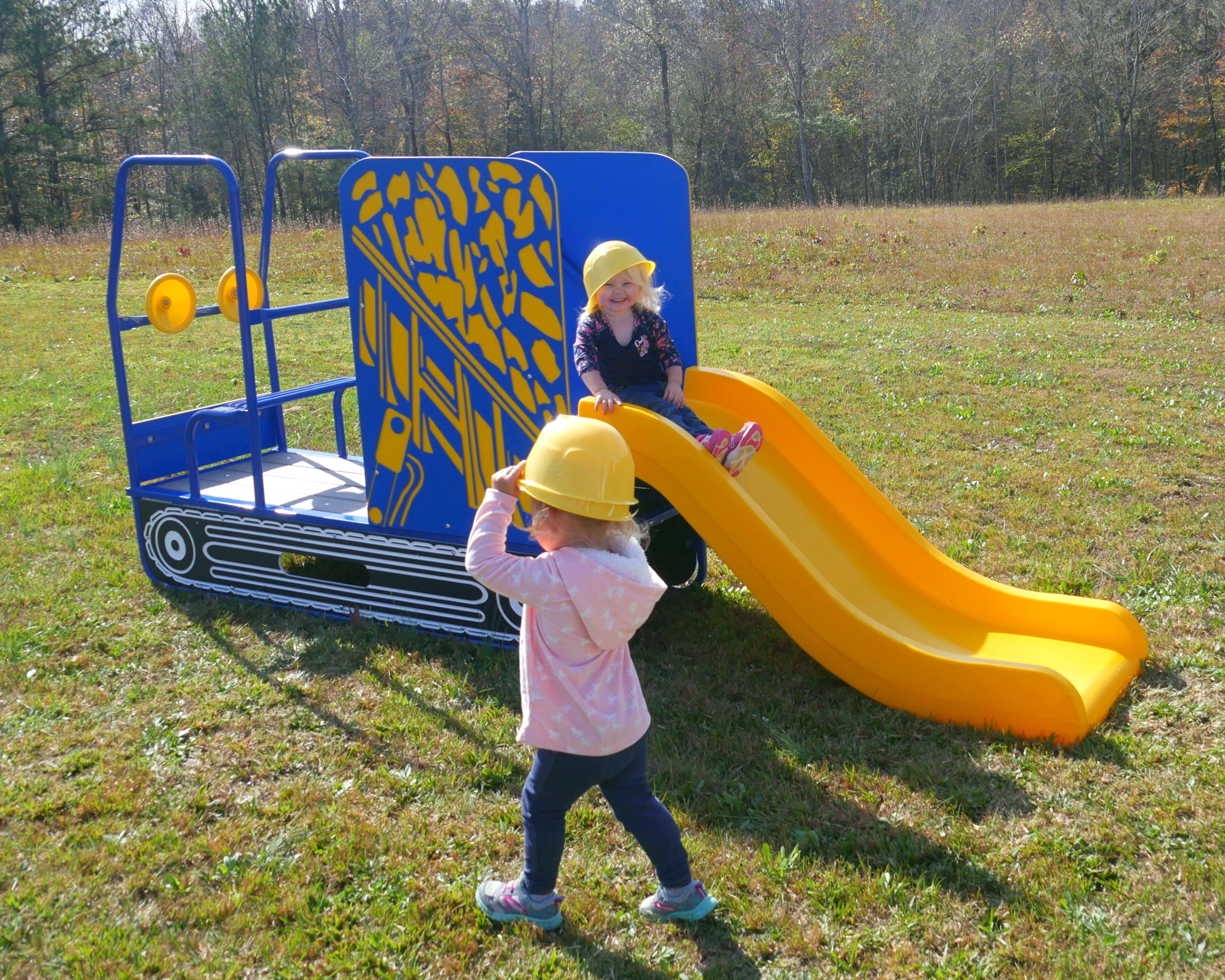 Bright yellow and blue dump truck-themed playground slide with two toddlers wearing yellow construction hats on grass