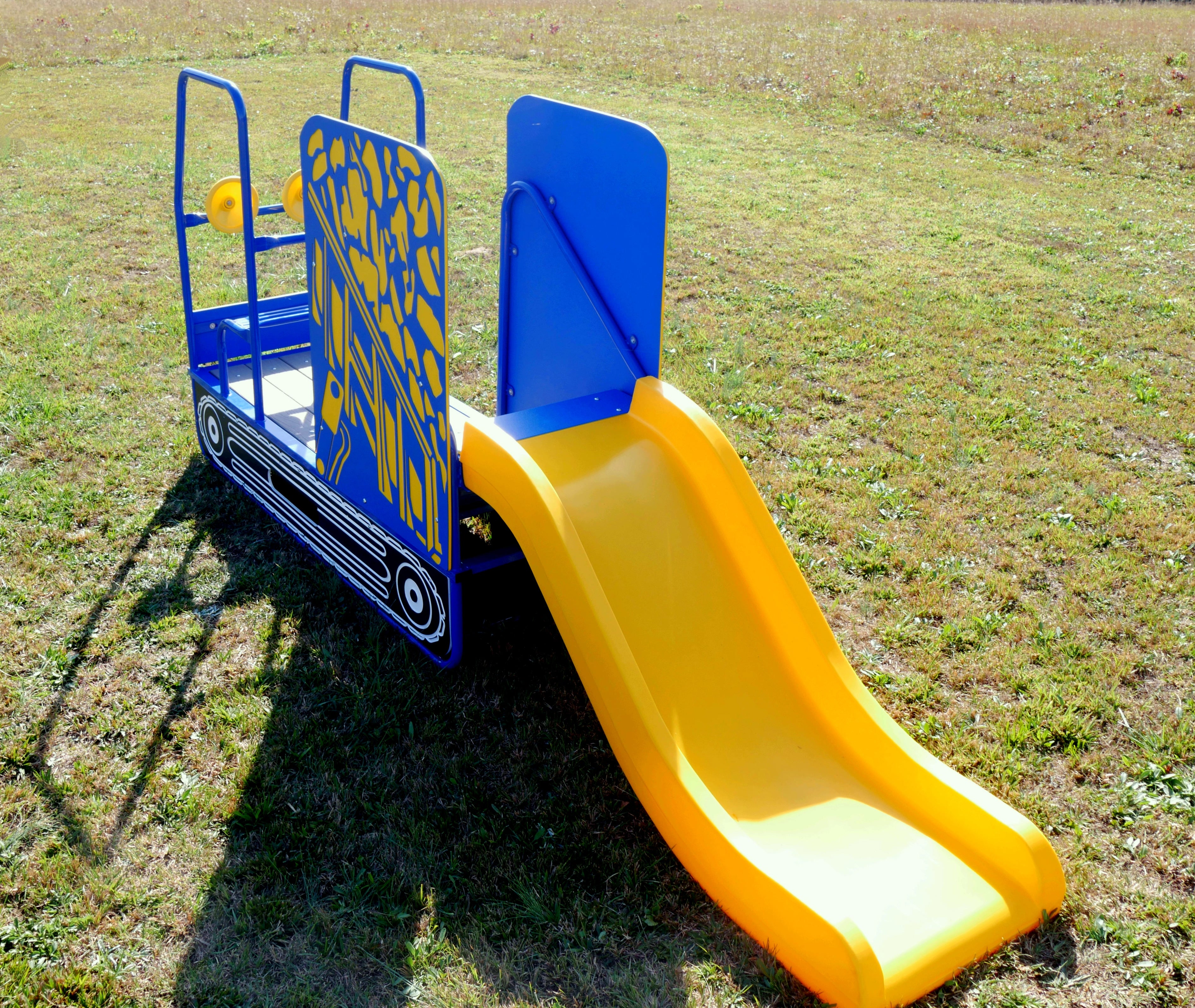 Compact blue and yellow dump truck-themed playground slide with twin steering wheels, climbing steps, and front bench on grass