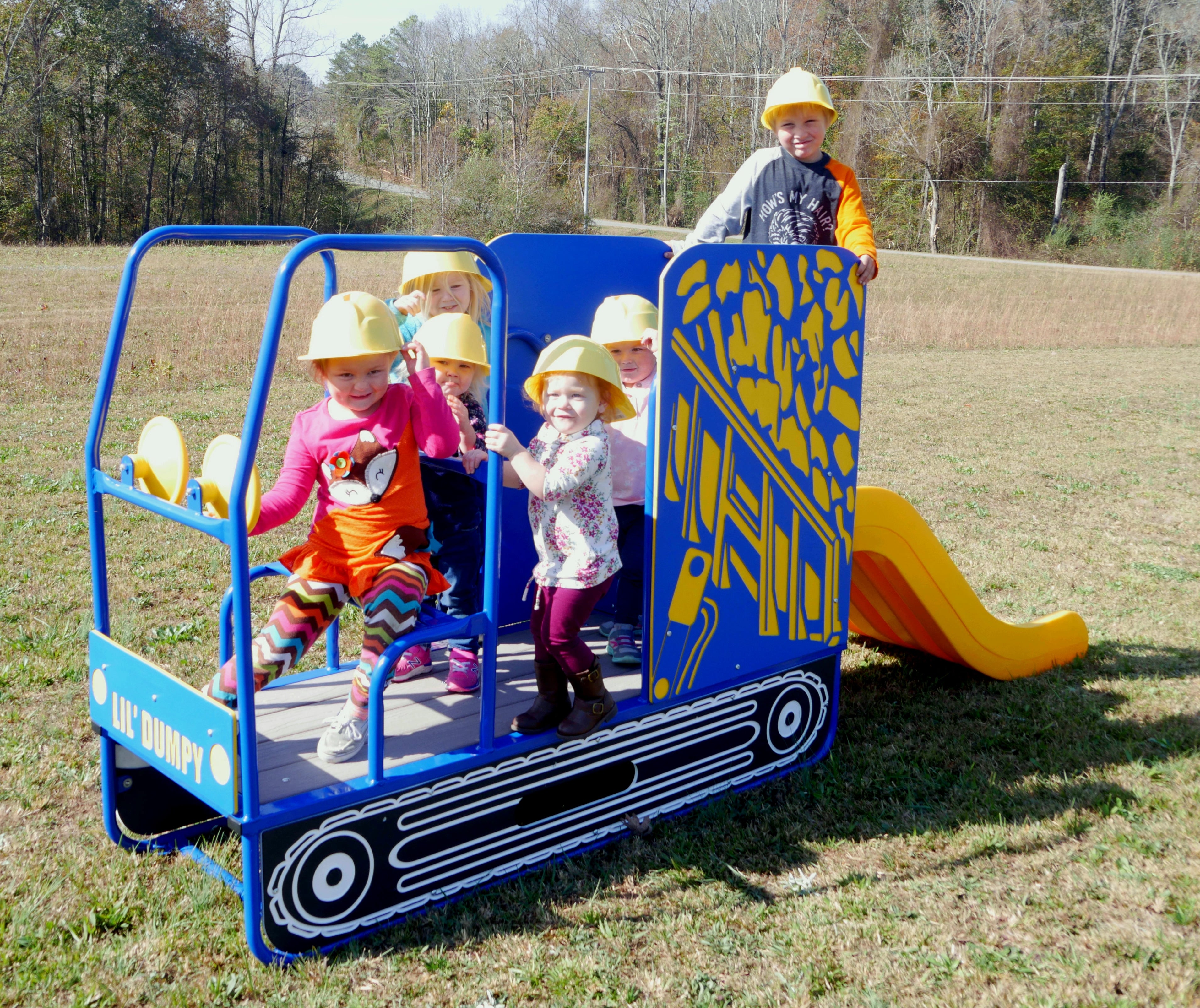 Children wearing yellow construction hats playing on a blue and yellow dump-truck themed playground slide with twin steering wheels outdoors