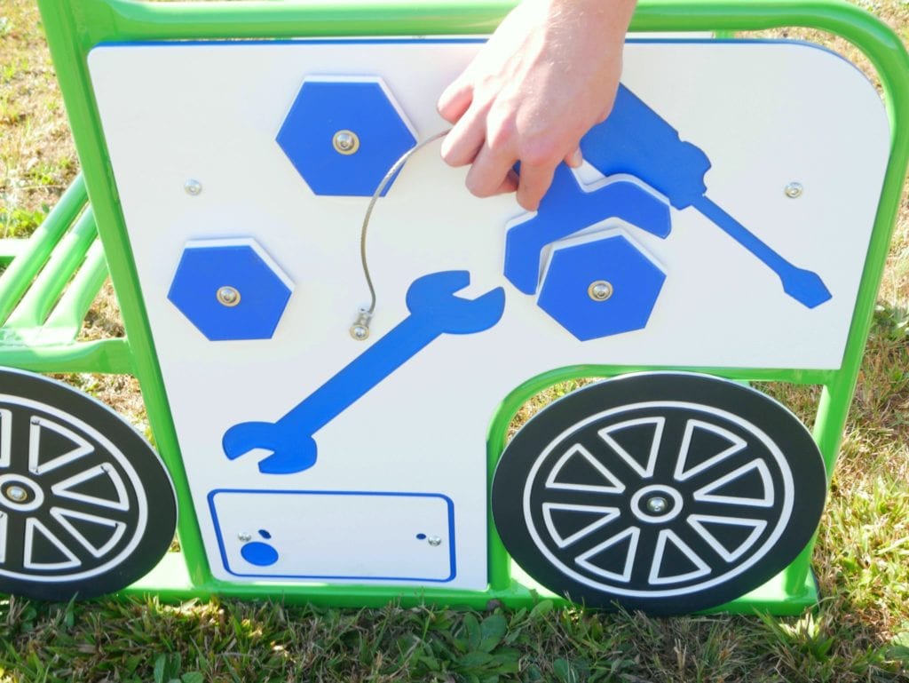 Green outdoor play panel with blue tool shapes and rotating hexagonal knobs, child’s hand engaging tactile features