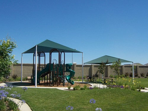 Green triangular shade structure with three posts covering playground equipment and picnic area in a park setting