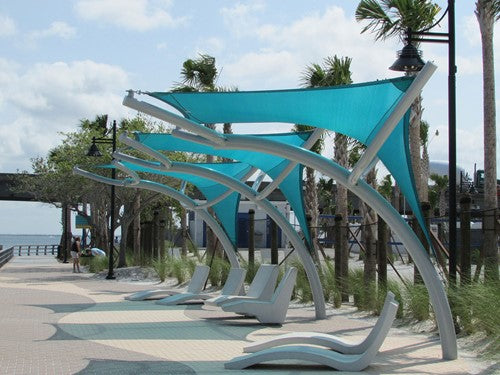 Turquoise fabric shade structures with curved metal supports over lounge chairs along a waterfront promenade