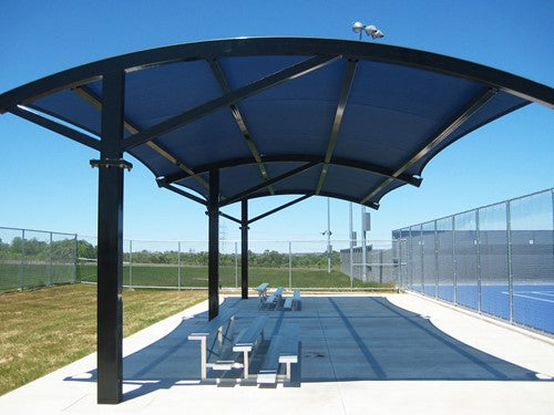 Arched cantilever shade structure with blue fabric roof over outdoor bleachers beside tennis courts