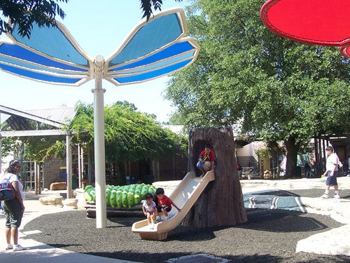 Butterfly wings shade structure with blue fabric panels over playground slide and climbing features in outdoor park setting