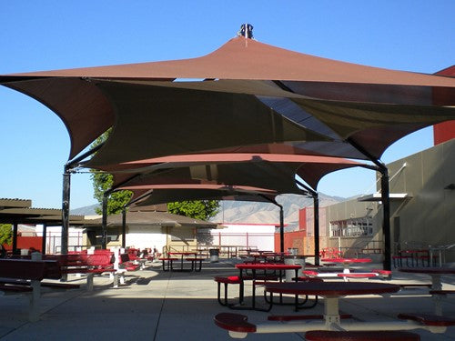 Mariner Pyramid Shade Structure with overlapping upper and lower fabric layers and four posts over outdoor picnic tables in a school courtyard